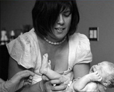 Une femme porte un chandail blanc et un collier de perle. Dans ses bras, elle tient un bébé naissant qui semble pleurer. Une autre paire de mains gantée est dirigée vers le bébé. Le bébé est nu. La photo est en noir et blanc.