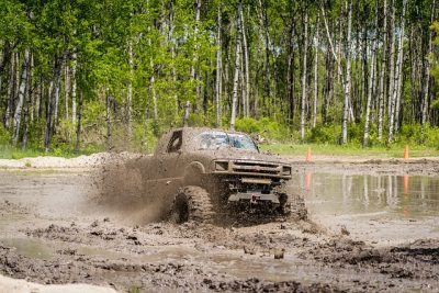 Un camion deux portes avec de grosses roues est dans un trou de boue. Il est complètement couvert de terre. Derrière le trou de boue, une grande forêt.