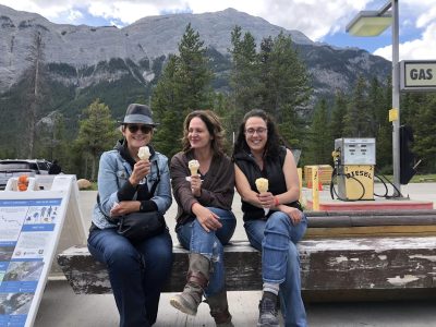 Trois dames sont assises sur un banc de bois foncé. Elles mangent de la crème glacée. Derrière elles, il y a une pompe à essence, des sapins et de grandes montagnes. Les trois dames sourient.
