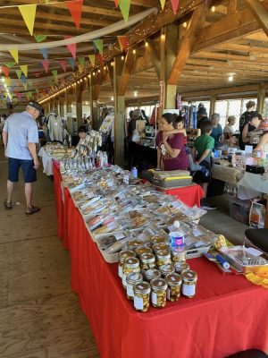 Une table napée rouge est couverte de produits à vendre (ail confite, pain, paniers divers). Plusieurs tables du genre sont alignées en arrière plan. Une dame et son enfant sont derrière la table. Un client regarde les produits. Plusieurs clients sont en arrières plans. Le tout est situé dans ce qui semble être une grange avec un toit en bois.