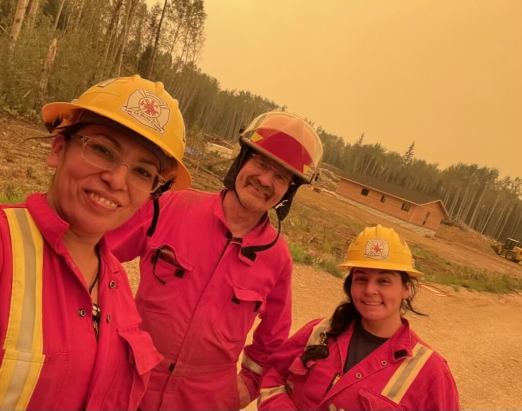 Trois personnes prennent un selfie. Ils sont habillés de combinaisons rouge et portent des casque jaunes de pompier. Ils sont à l'extérieur devant une forêt.