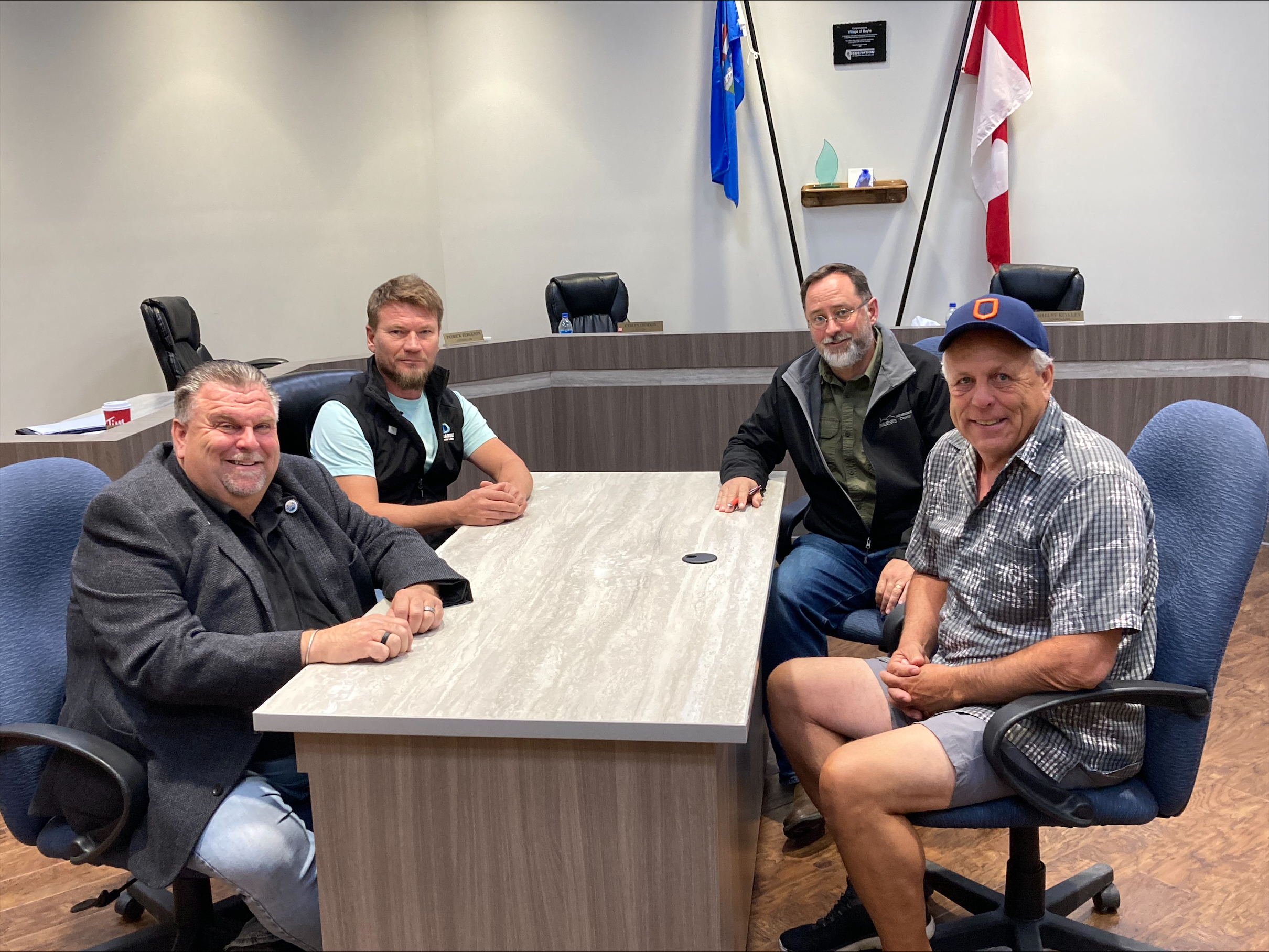 Quatre hommes sont assis autour d'une table et sourient à la caméra. La table est dans le bureau du conseil municipal. Sur le mur, il y a le drapeau canadien et le drapeau albertain.