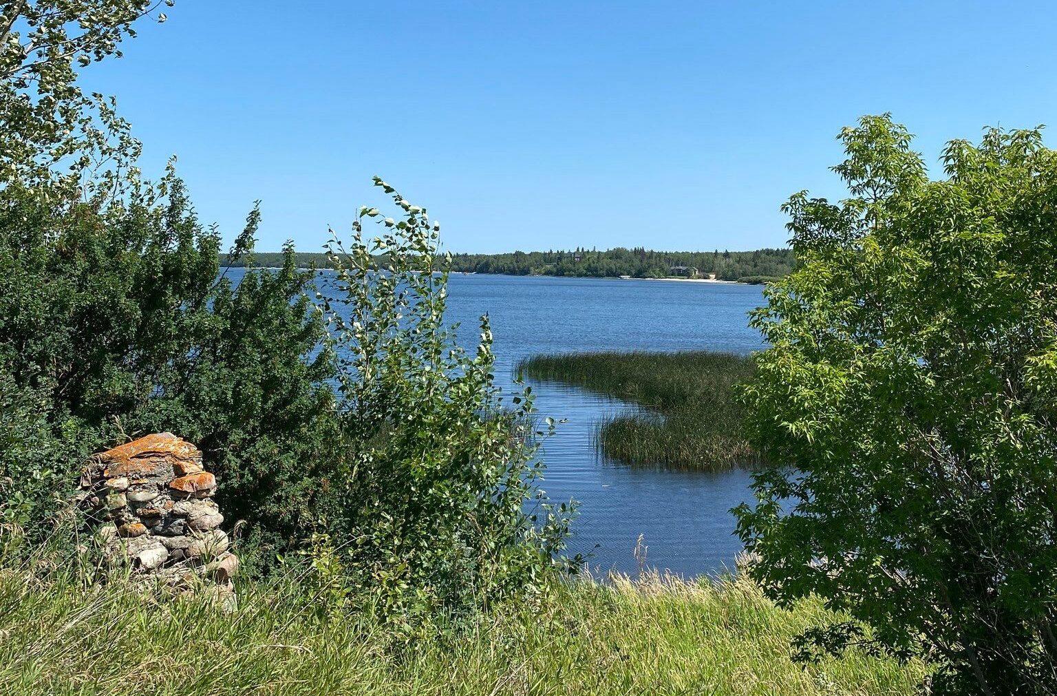 Un lac bleu précédé d'un bout de terre avec quelques arbustes et des pierre. Au loin, on voit l'autre rivage, avec de la végétation.