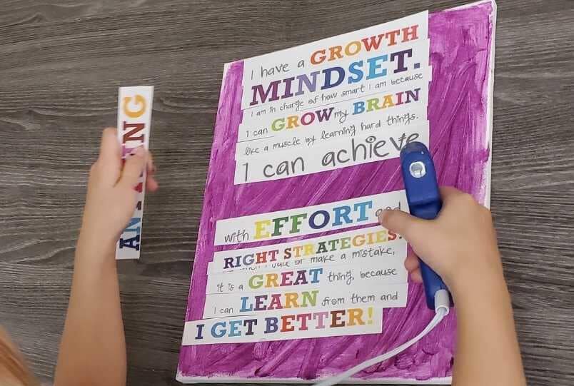 Un cahier mauve sur lequel est inscrit « I have a growth mindset » ainsi que plusieurs phrases encourageantes est tenues par de petites mains d'enfants. L'enfant tient un mot dans une main et un bâton de colle chaude dans l'autre.