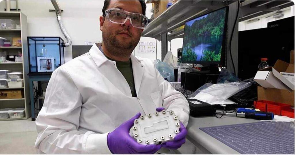 Un homme dans un laboratoire porte un sarrau blanc et des lunettes protectrice. Il présente à la caméra un petit objet de plastique blanc.