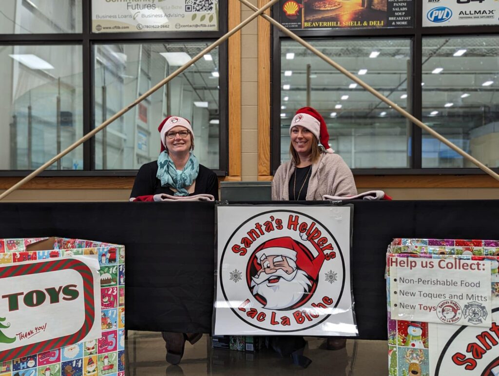 Deux femmes portent des chapeaux de Père Noël. Elles sont assises à une table décorée d'une affiche « Santa's Helpers »